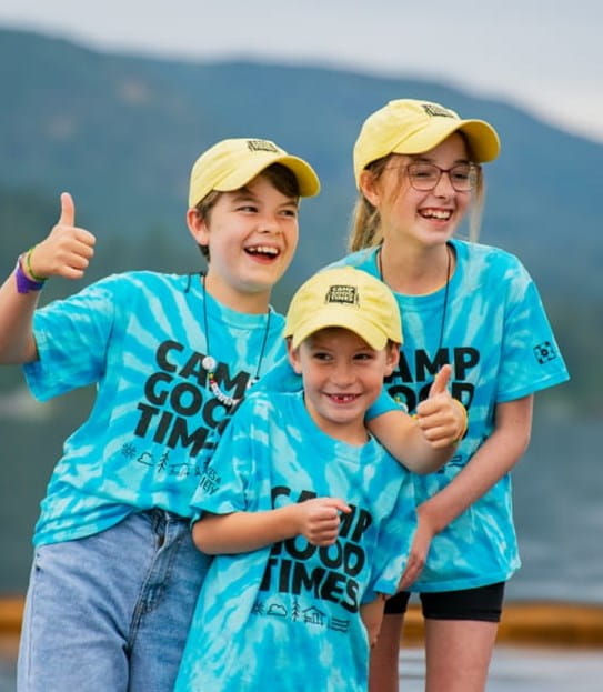 Three kids wearing matching Camp Goodtimes shirts and hats and smiling.