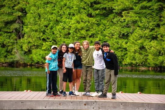 A group of campers posing on the dock in front of trees with their arms around each other’s shoulders.