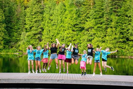 A group of campers holding hands and jumping on the dock in front of trees.