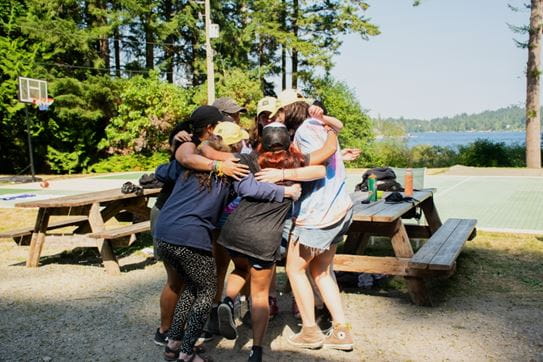 A group of campers holding each other in a hug in front of picnic tables.