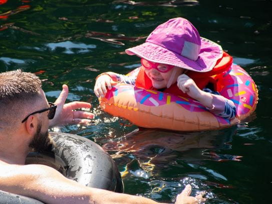 A man in the lake holding his arms open to a smiling toddler wearing a floating device, goggles and a hat.