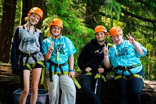 A group of four kids wearing helmets and harnesses, smiling and holding up peace signs in front of trees.