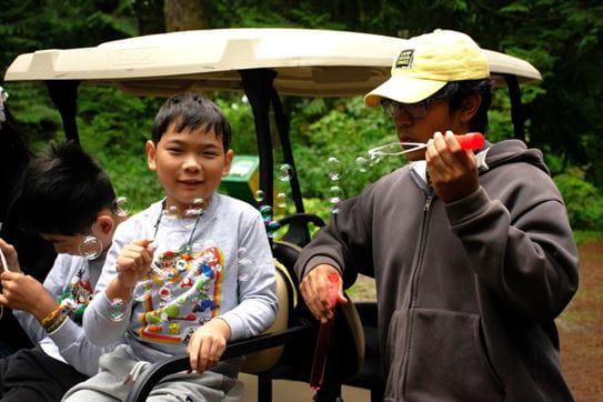 Two kids blowing bubbles on the back of a golf cart.