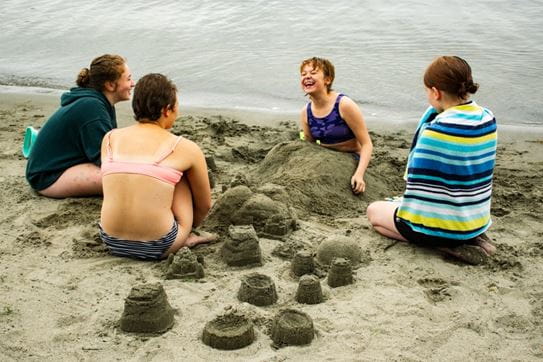 Four campers playing in the sand by the water beside sandcastles.