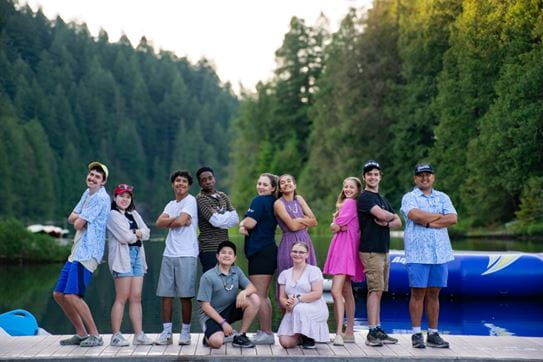 A group of campers posing on a dock in front of trees with their backs to each other.