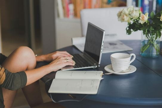 A person sitting at a table and typing on their laptop. A teacup, vase with flowers and date book are also on the table.