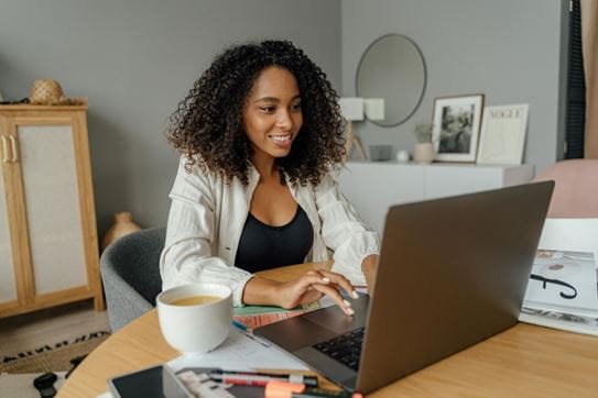 A person sitting in their living room at a table typing on their laptop and smiling.