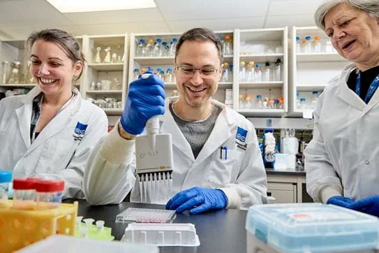 Trois chercheurs à l’œuvre dans un laboratoire sourient. Leur sarrau arbore le logo de l’Institut de recherche en immunologie et en cancérologie (IRIC).