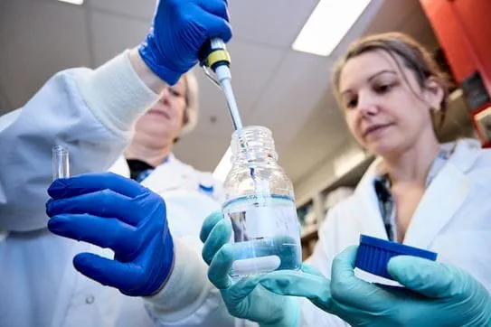 Deux chercheuses portent un sarrau et des gants; l’une tient un flacon de verre contenant un liquide clair et l’autre plonge un outil dedans.