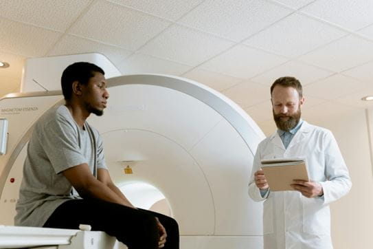 Un homme assis sur une table d’imagerie par résonance magnétique. Un médecin se tient à ses côtés, penché sur ses notes.