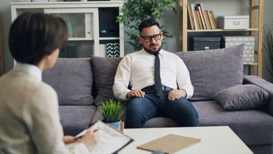 A man sitting on a couch, speaking to a person holding a clipboard.
