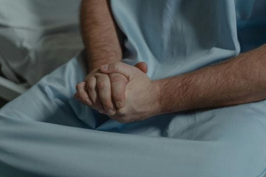 Gros plan d’une personne en blouse d’hôpital, assise sur un lit, les mains jointes posées sur les genoux.