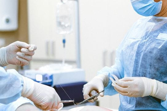 Two surgeons dressed in surgical gowns stitching a person with a pair of scissors and surgical thread.