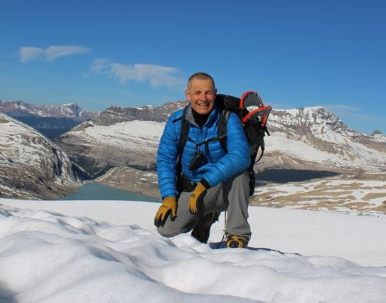 Ron Luciuk smiling and squatting on the snow surrounded by mountains.