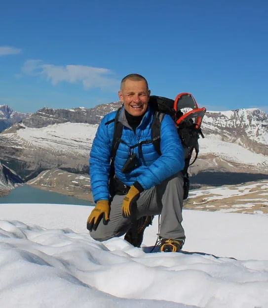 Ron Luciuk smiling and squatting on the snow surrounded by mountains.