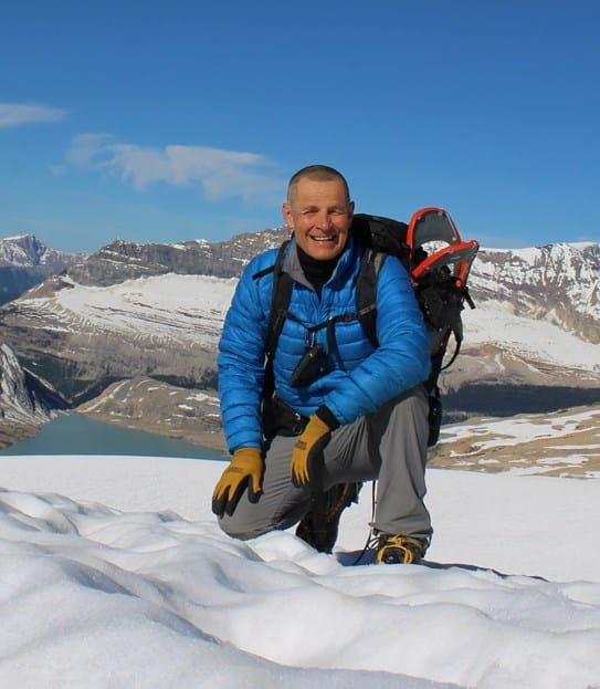 Ron Luciuk souriant, accroupi dans la neige dans un paysage montagneux.