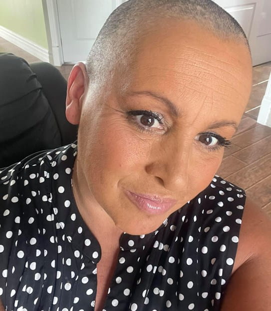 Annie Lemieux, with a shaved head, sitting at home, smiling.