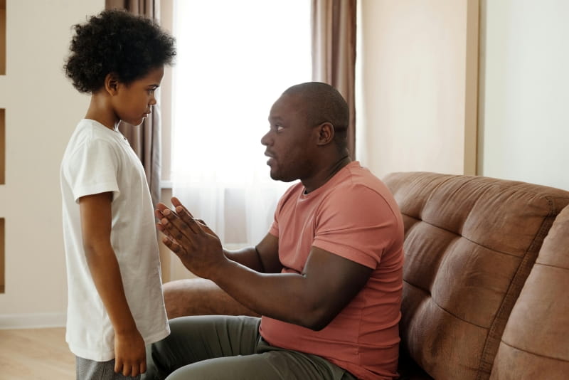 A father sitting on a couch and talking to his son standing in front of him.