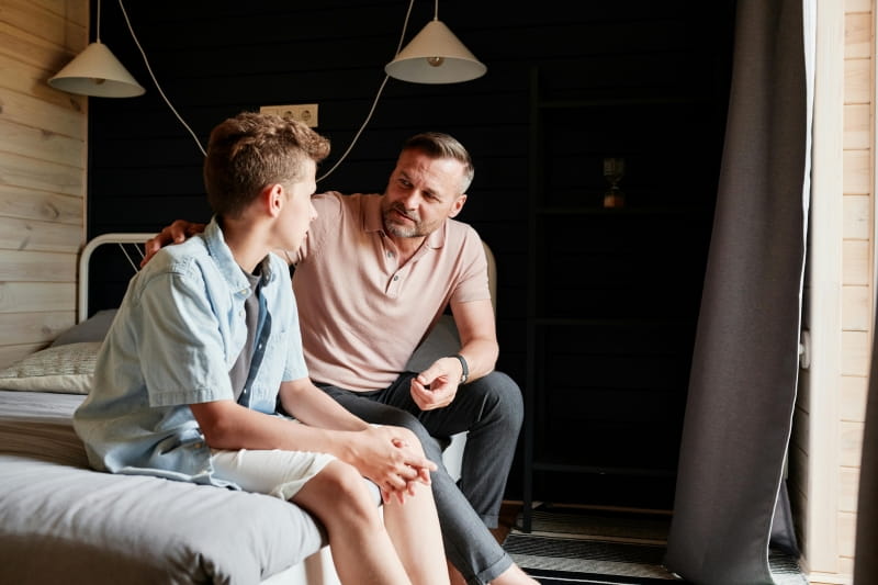 A father and son sitting on a bed. The father’s hand is on his son’s back.