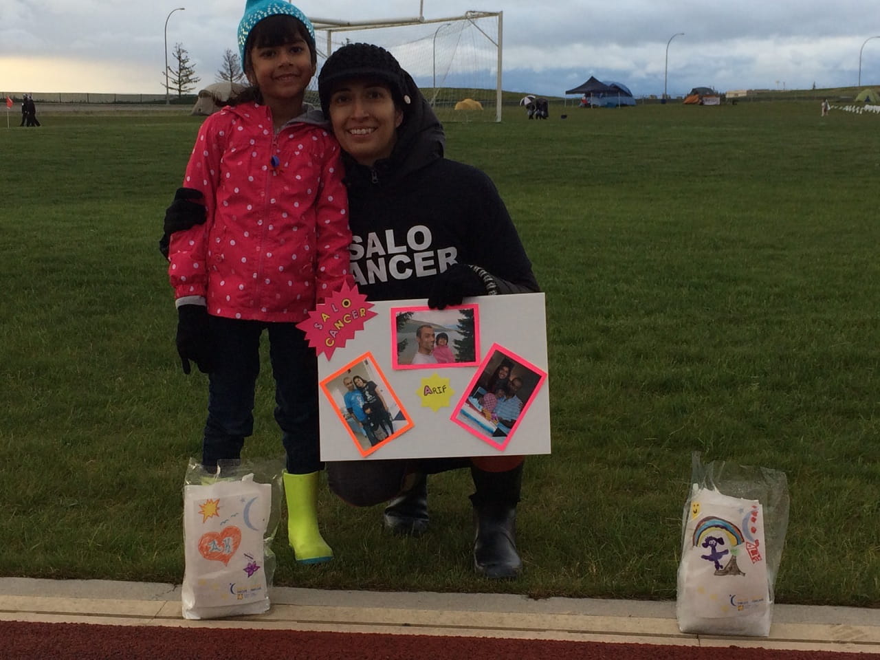 Zahra Khakoo is kneeling, holding her daughter and a sign with photos of her husband Arif. There are two luminaries on a bench in front of them. 