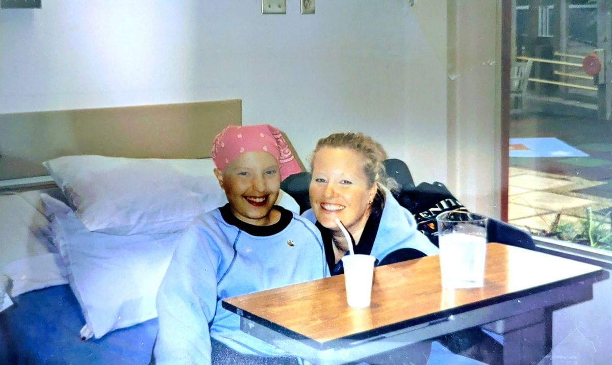 Kyla est assise dans un lit d’hôpital et l’une de ses ses infirmières se tient à ses côtés. Les deux sourient.