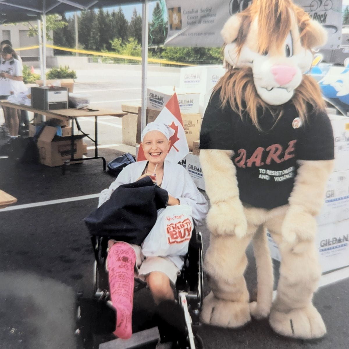 Kyla is in a wheelchair with her leg in a cast, a tour de rock mascot is next to her wearing a lion costume.