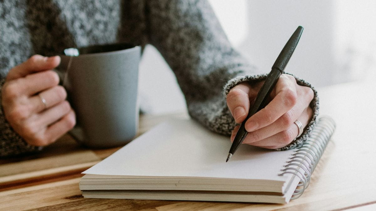 Close-up of a person writing in a notebook with a pen while holding a mug. They are seated at a wooden table. 