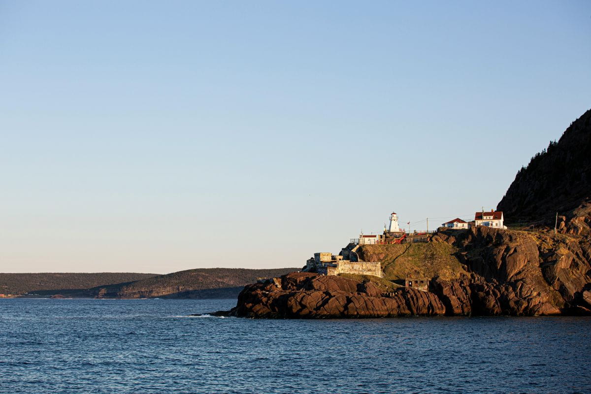 A lighthouse sits on a rocky cliff beside calm blue water under a clear sky in St. John's, NL. 