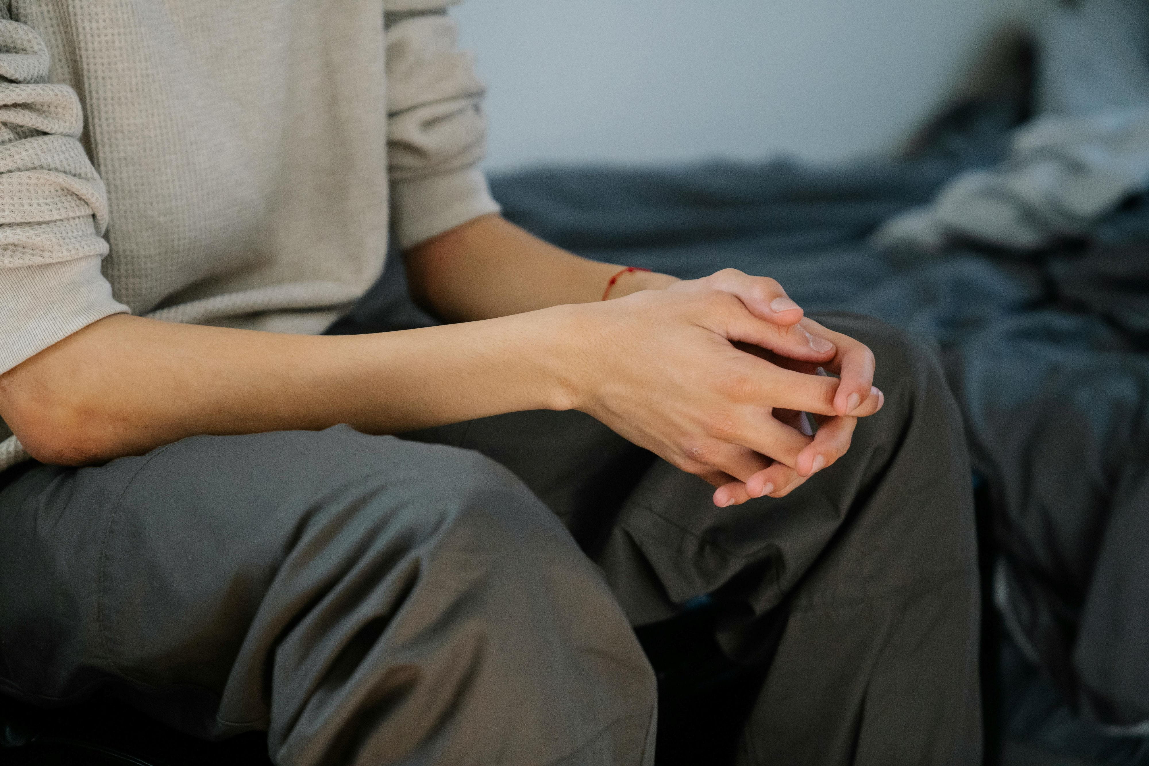 Une personne est assise sur le bord d’un lit, les mains jointes.