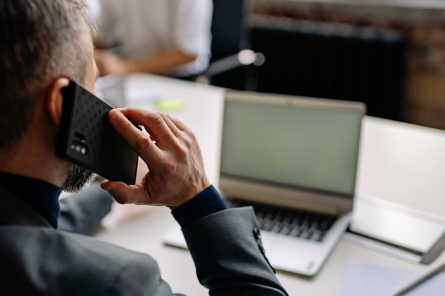 Une personne assise devant un portable qui tient un téléphone cellulaire contre son oreille.