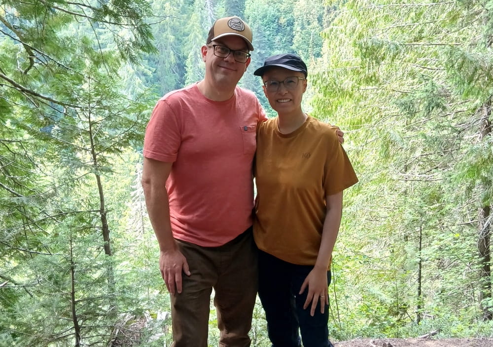 Rita and her husband standing in a forest.