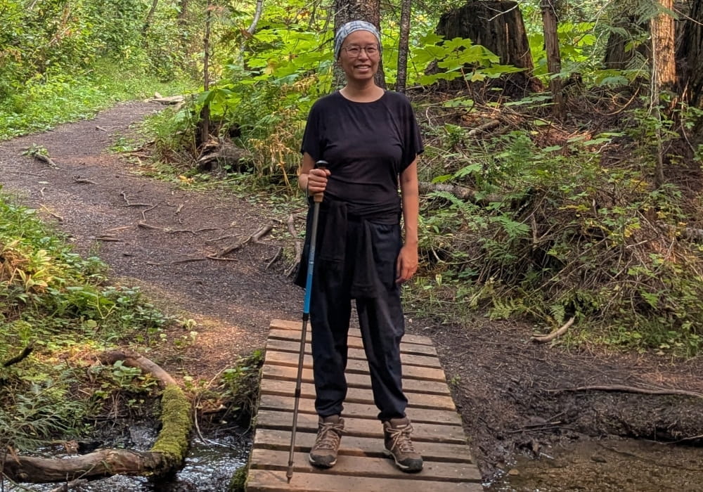 Rita standing in a forest holding a walking stick and smiling.