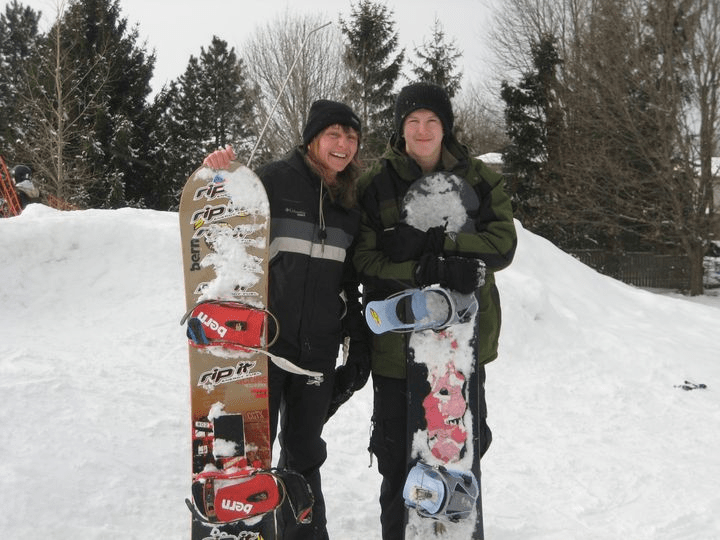 Tanja and Nick are standing with their snowboards in front a small snow hill and trees. 