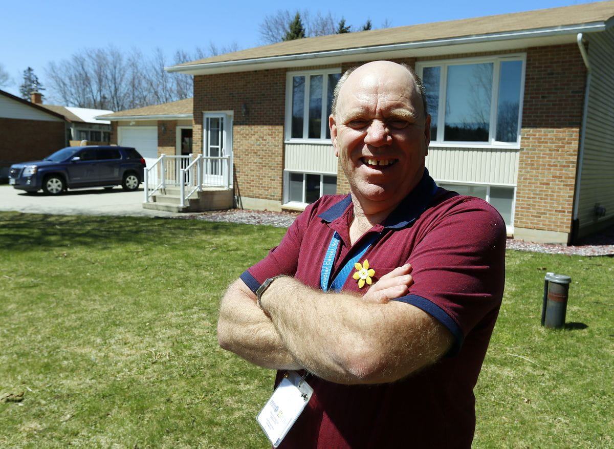 Souriant, les bras croisés, Wayne se tient devant chez lui, portant une épinglette jonquille et un cordon au cou.