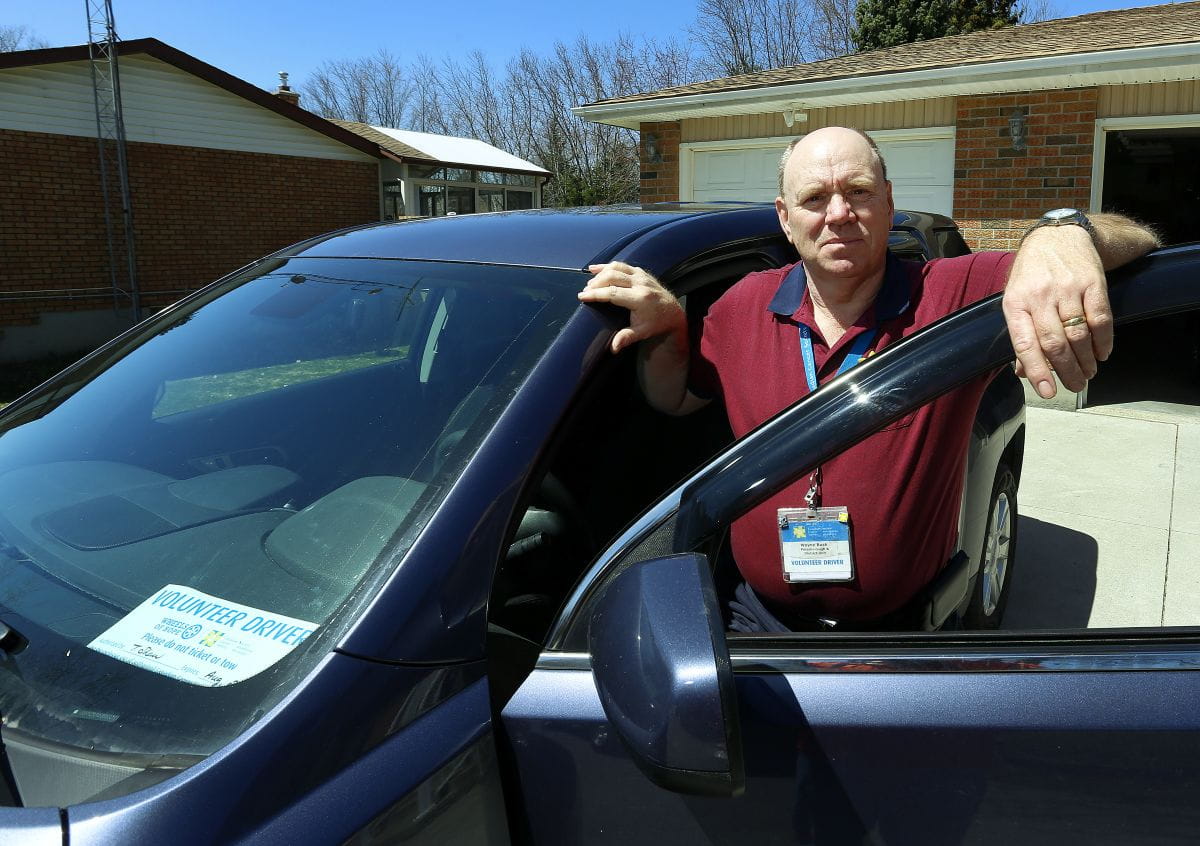 Wayne à l’extérieur de sa voiture, une pancarte de chauffeur bénévole dans le pare-brise. 