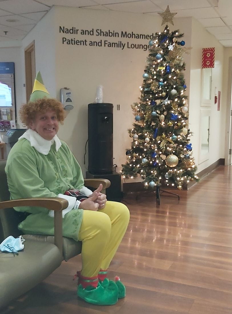 Terry sits in front of a Christmas tree in the hospital. He is dressed in an elf costume. 