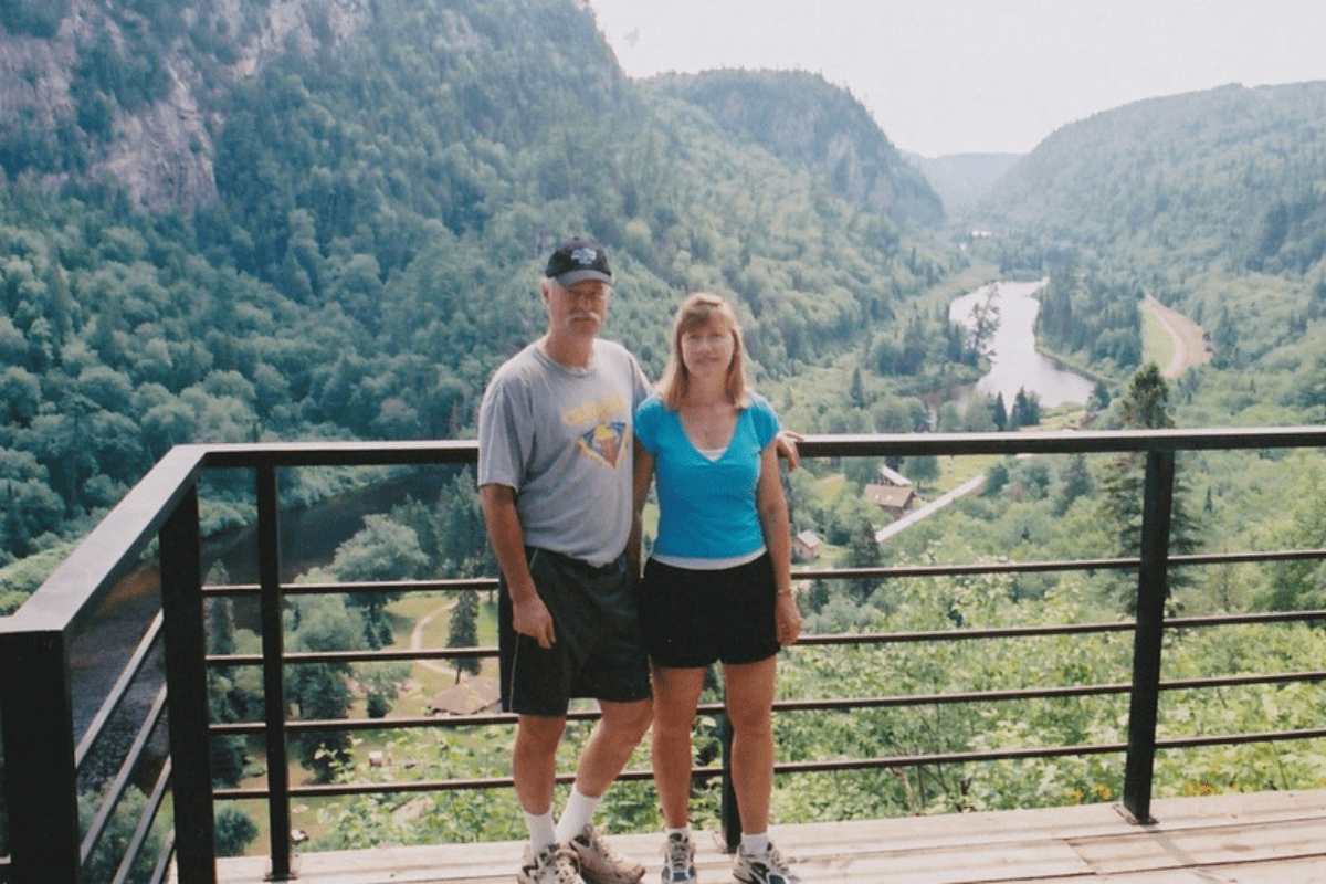 Stuart and Elaine stand on a deck overlooking a green mountain valley with a river below. 