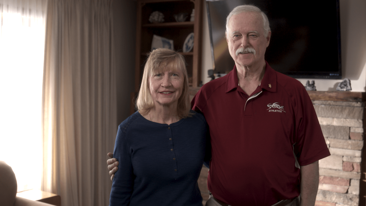 Stuart and Elaine stand in their living room. They have their arms around one another and smile. 