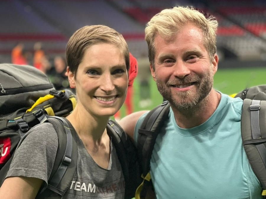 Catherine et son meilleur ami Craig sourient dans un stade, les bras l’un autour de l’autre, chacun avec un sac à dos.