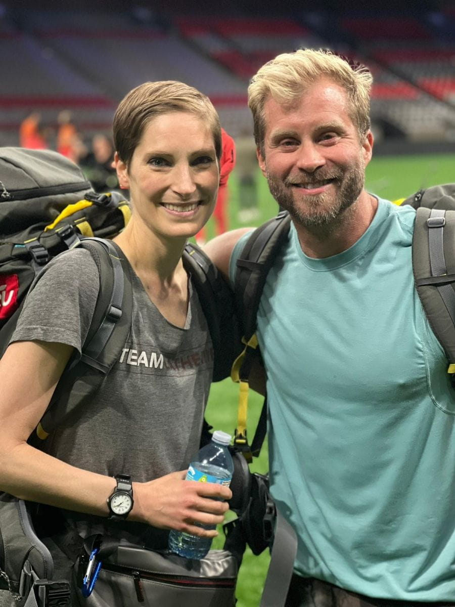Catherine and her best friend Craig are standing in a stadium and smiling with their arms around each other. They are both wearing backpacks. 