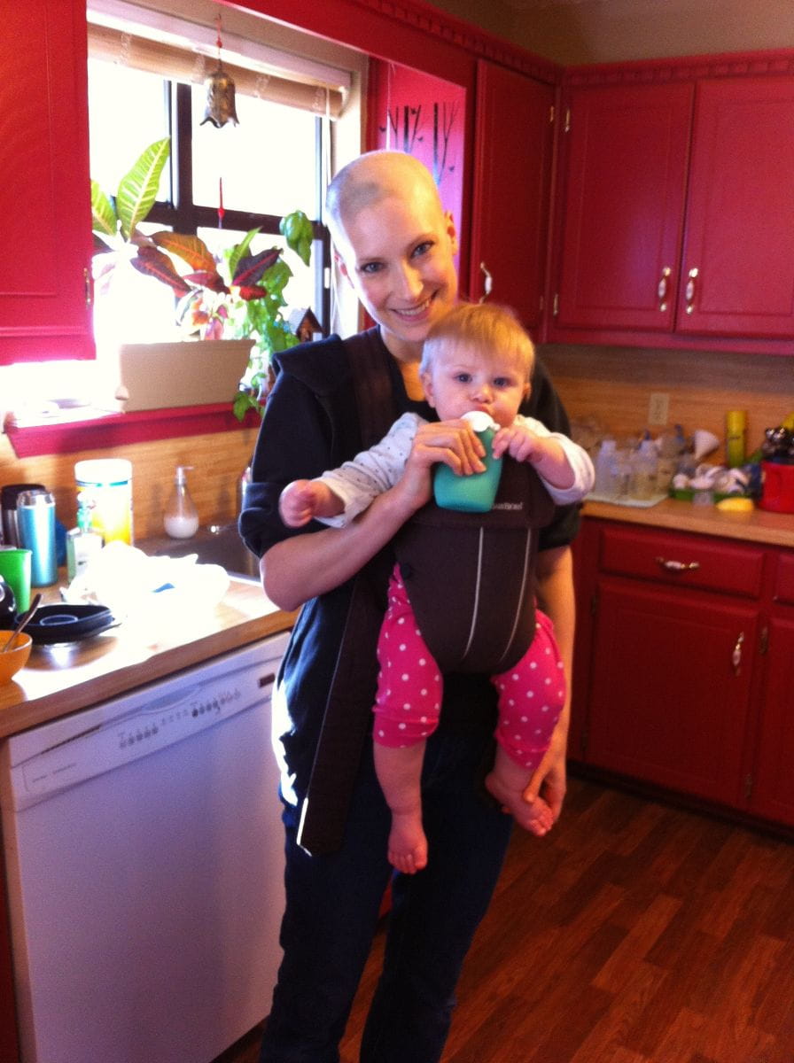 Catherine stands in her kitchen and feeds her daughter who is in a baby carrier. 
