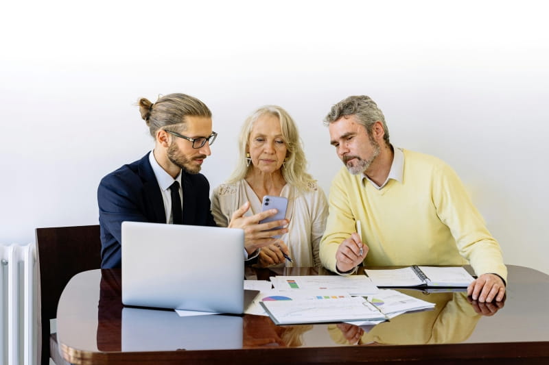 Three people sitting at a table in front of a laptop and papers. One person shows the other 2 people something on his phone.