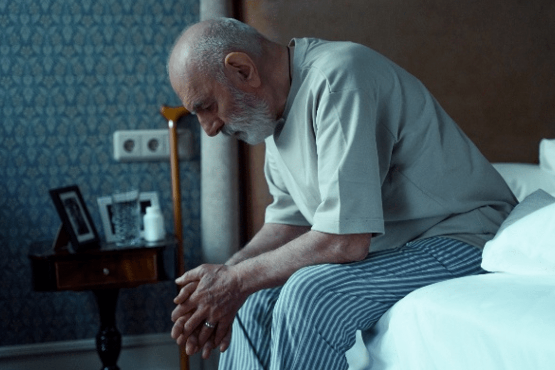 A man sitting on the edge of a bed and leaning on his knees with his forearms.