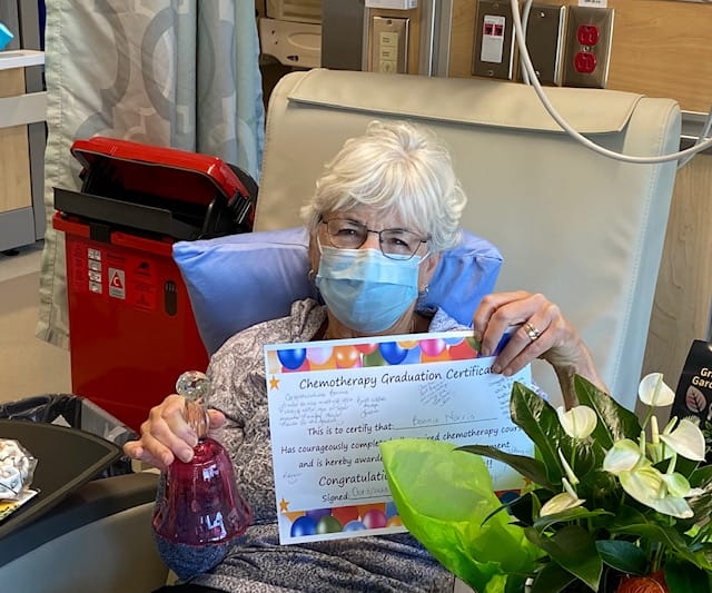 Bonnie est assise sur une chaise à l’hôpital, tenant son certificat de fin de chimiothérapie. Elle porte un masque et tient une clochette dans son autre main. Il y a un bouquet de fleurs à côté d’elle.