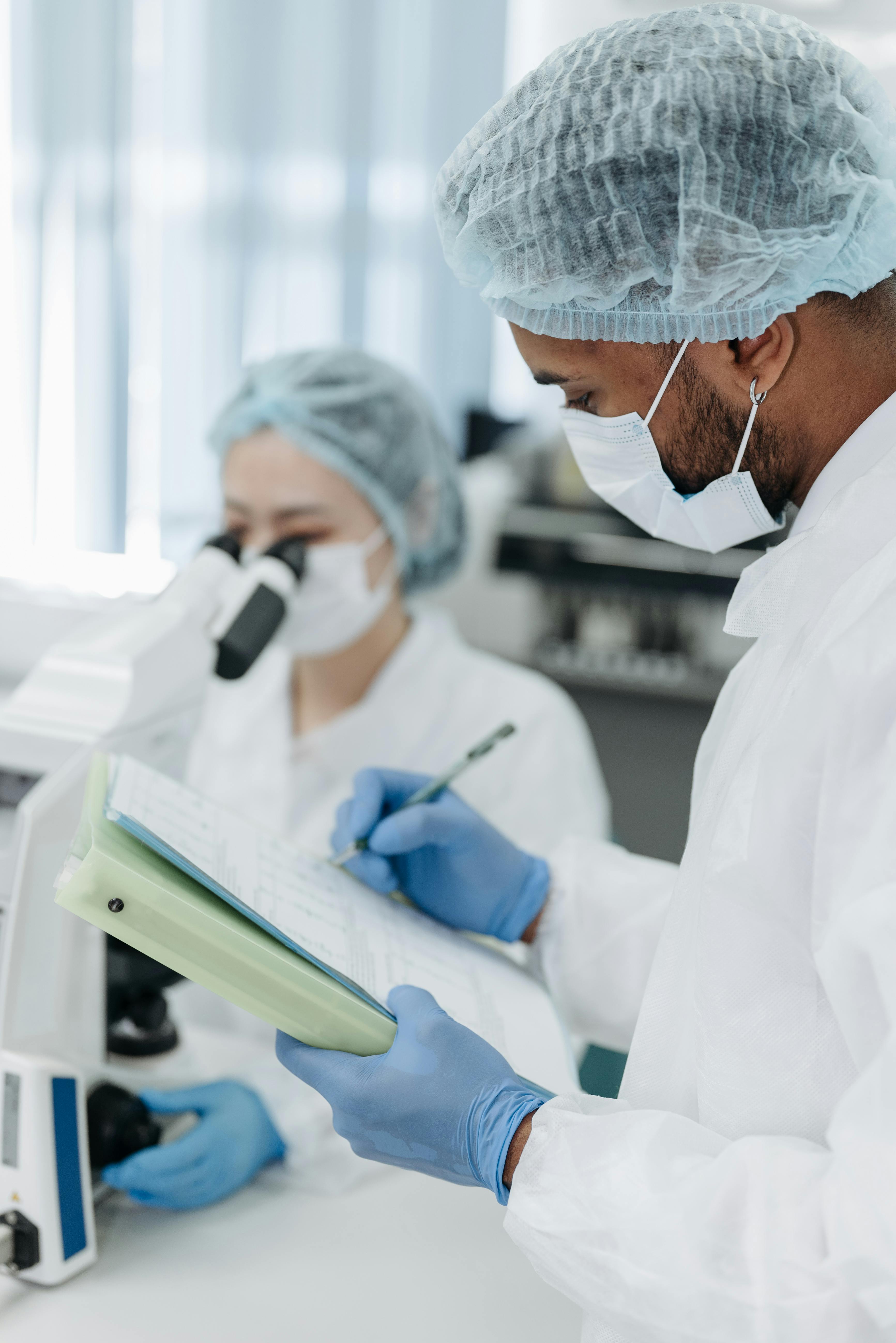 Two people wearing lab coats, face masks and hair coverings. One looks into a microscope and the other writes in a notebook.