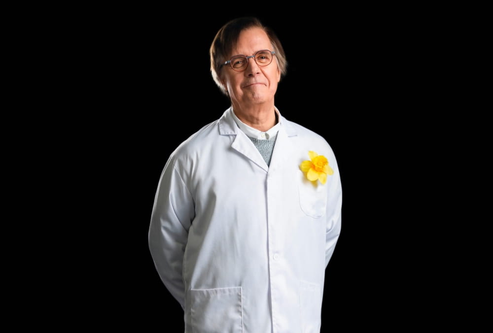 Claude Perreault, Ph.D., souriant devant l’appareil photo, les mains dans le dos, vêtu d’un sarrau arborant une épinglette jonquille.