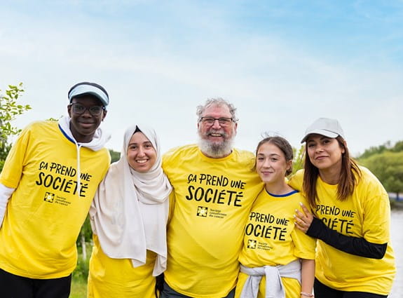 Un groupe de personnes portant des T-shirts « Il faut une société » se tient devant une zone boisée et sourit.