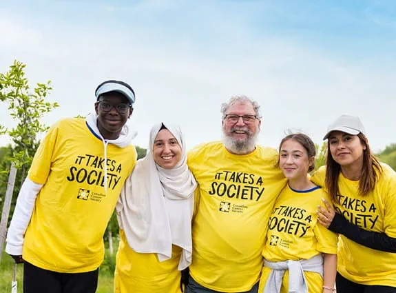 A group of people wearing ‘It takes a society” T-shirts are standing in front of a wooded area. They are laughing and smiling.