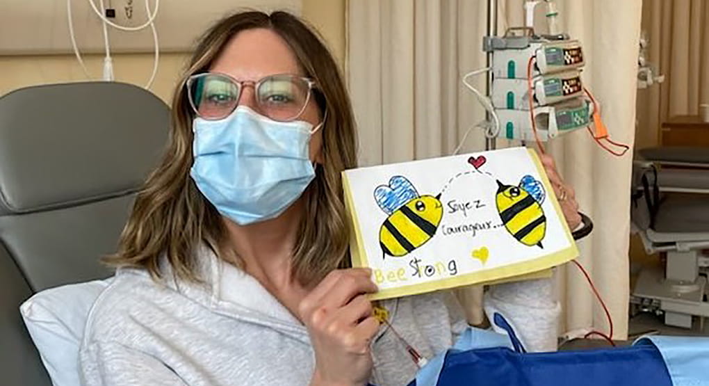 Amélie sitting in a hospital bed and is showing a drawing with the words “be strong”