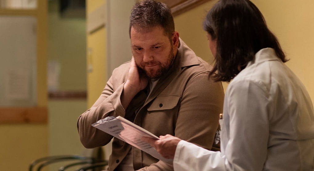 A doctor is holding a medical chart and talking to a person as they sit in a hospital hallway.